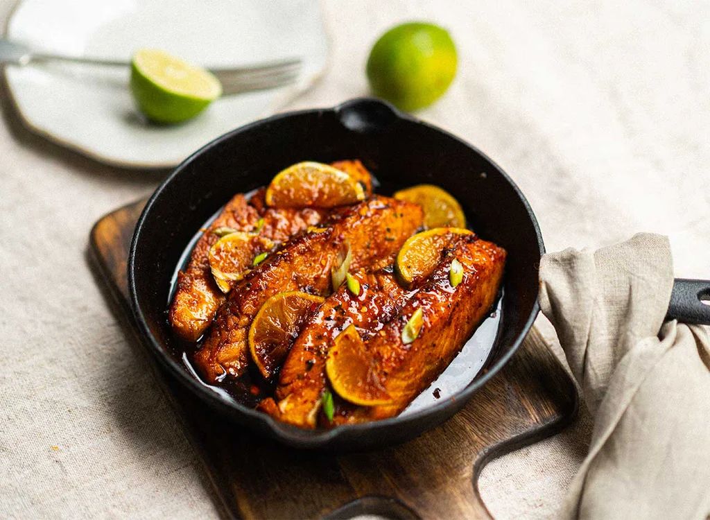 Fish dish with citrus slices in a black skillet on a wooden board, with limes in the background.